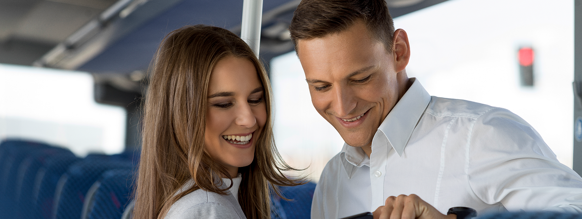 Two smiling people on a bus looking at a smartphone.